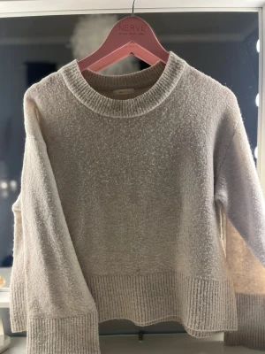 Beige stickad tröja med breda muddar - Mysig beige stickad tröja med rund halsringning och breda ribbstickade muddar vid ärmslut och nederkant. Tröjan har en avslappnad passform och är tillverkad i ett mjukt material som passar perfekt för kyligare dagar.