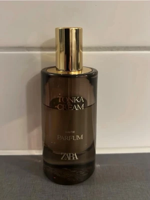 Zara Tonka Cream Eau de Parfum - Tonka Cream Eau de Parfum från Zara i en elegant, mörkbrun glasflaska med guldfärgat lock. Flaskan har en rundad form och stilren design med text i guld. Perfekt för dig som gillar lyxiga och moderna dofter.