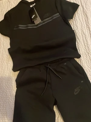 Svart set från Nike med logga - Snyggt svart set från Nike med t-shirt och shorts. T-shirten har diskret Nike-logga och en tunn grå rand över bröstet. Shortsen har dragsko i midjan och fickor samt en matchande logga. Perfekt för en sportig och avslappnad stil.
