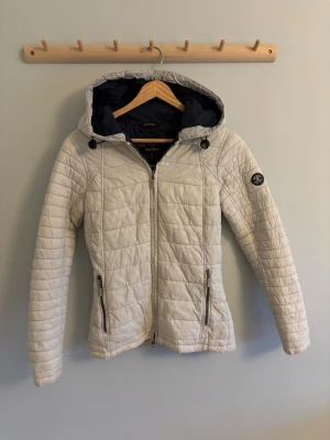 Ljusbeige pufferjacka med huva - Snygg ljusbeige pufferjacka från barbour med quiltat mönster, dragkedja framtill och två fickor med dragkedja. Jackan har en justerbar huva med snörning och ett diskret märke på ärmen. Perfekt för kalla vinterdagar och riktigt skön passform. Har några svarta fläckar på baksidan