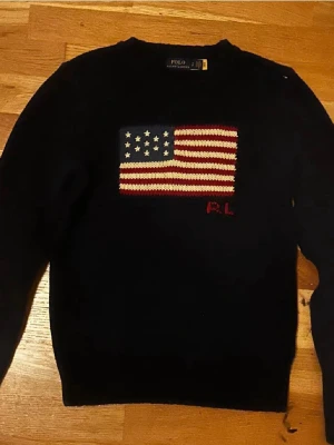 Ralph Lauren flagg tröja - Blå stickad tröja från Polo Ralph Lauren med amerikansk flagga i rött, vitt och blått på bröstet samt RL-broderi. Den är lite krympt men fortfarande väldigt skön och snygg! Tröjan har rund halsringning, långa ärmar och ribbstickade muddar. Klassisk och tidlös design. Det är bara o höra av dig vid minsta fråga!          