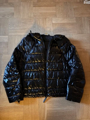 Svart glansig ZARA jacka  - Säljer en svart, glansig pufferjacka med huva och dragkedja framtill. 