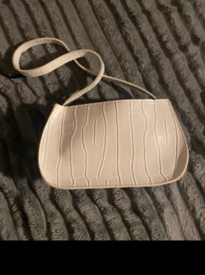 Beige/vit axelremsväska i krokopräglat skinn - Stilren beige/vit handväska med axelrem och krokodilpräglat mönster. Väskan har en mjukt rundad form. Perfekt för dig som gillar trendiga accessoarer med lite extra attityd.