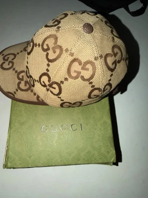  Gucci Jumbo - Säljer en beige keps från Gucci med det klassiska GG-monogrammet i brunt över hela kepsen. Kepsen har böjd skärm och justerbart spänne bak i brunt läder. Materialet är canvas med detaljer i läder. Perfekt för dig som vill ha en lyxig och trendig accessoar. Kepsen har inga skador. Box medföljs dessutom har kepsen ett chipp som äkthetmaterial. 