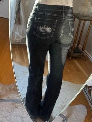 Lågmidjade jeans med tryck på fickan - Säljer ett par svarta lågmidjade bootcut jeans med krona broderad på bakfickorna. Jeansen har vit kontrastsöm, klassisk femficksmodell och är tillverkade i stretchigt denim för skön passform.