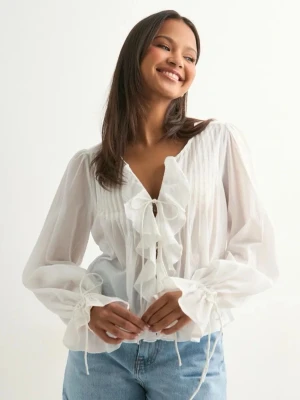 Nelly - Stunning Flounce Blouse - Söt vit blus med volangdetaljer framtill och knytning både vid halsringningen och vid ärmsluten. Blusen har en luftig och lätt transparent känsla, samt långa ärmar med fina rynkade detaljer. Perfekt att styla med jeans för en trendig look. Alla etiketter och taggar sitter kvar, blusen har aldrig använts. Original pris 399 kr.
