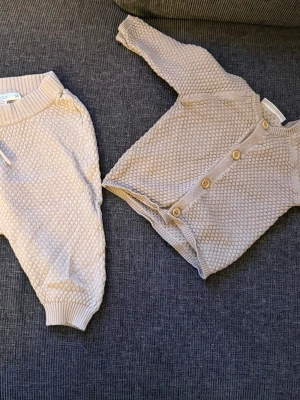 Beige stickat babyset från Ergee - Säljer ett gulligt stickat set för baby från Ergee i storlek 56. Setet består av en beige body med träknappar framtill och matchande byxor. Båda delarna har ett fint strukturmönster och mjuka ribbade muddar. Perfekt för mysiga dagar hemma.