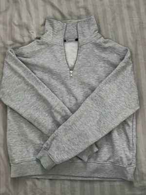 Ljusgrå halvzip hoodie - Snygg ljusgrå hoodie med halvzip och hög krage. Tillverkad i mjukt sweatshirtmaterial som känns skönt mot huden. Klassisk passform med ribbade muddar vid ärmslut och nederkant. Perfekt för en avslappnad och stilren look.