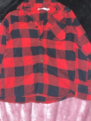 Röd och svart rutig skjorta - Klassisk röd och svart rutig skjorta med knappar framtill och en bröstficka. Skjortan har lång ärm och avslappnad passform, perfekt för en cool och avslappnad stil. Tillverkad i mjuk bomull som känns skön mot huden.