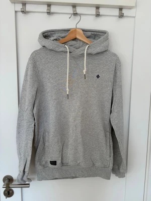 Grå hoodie med dragsko och brodyr - Snygg grå hoodie från Morris med vit dragsko i huvan och liten mörk broderad logga på bröstet. Klassisk passform med känguruficka framtill och ribbade muddar vid ärmslut och nederkant. Tillverkad i mjuk bomullsblandning för extra komfort.