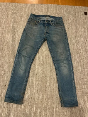 Nudie jeans - Säljer ett par snygga blå jeans från Nudie Jeans med raka ben och fem fickor. Jeansen har snygg slitning på låren och bakfickor med Nudie Jeans karakteristiska söm. Tillverkade i denim av bomull. Jag har växt ut de, ställ frågor vid minsta fundering😊👌
