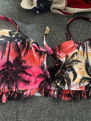 Färgglad bikiniöverdel med palmmönster - Säljer en bikiniöverdel med tropiskt palmmönster i rosa, orange och beige toner. Modellen har volangkant nedtill och en snygg knytning framtill. Perfekt för strandhäng och sommarvibbar!