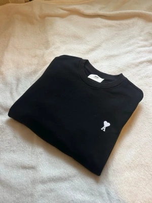Svart sweatshirt från Ami Paris - Svart sweatshirt från Ami Paris med rund halsringning och ikonisk vit broderad hjärtlogga med bokstaven A på bröstet. Tröjan har en klassisk passform och är tillverkad i mjuk bomull som känns skön mot huden.