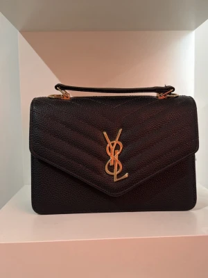 Svart YSL handväska i läder - Säljer en svart handväska i strukturerat läder med guldfärgade detaljer och ikoniska YSL-loggan framtill. Väskan har ett topphandtag och elegant, stilren design med kuvertformad lock. Väskan är i nyskick endast använt 1 gång.