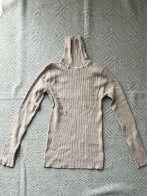 Beige ribbad polotröja - Säljer en beige ribbad polotröja med lång ärm och hög krage. Tröjan är figurnära och har en mjuk, stretchig känsla. Perfekt för lager på lager och enkel att matcha med olika stilar.