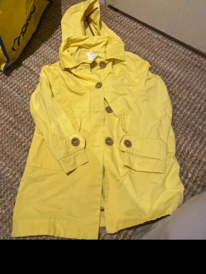 Gul regnjacka från Zara med huva - Snygg gul regnjacka från Zara med stora bruna knappar och praktisk huva. Jackan har två fickor framtill och detaljer vid ärmsluten. Perfekt för regniga dagar och ger en färgklick till din outfit. Tillverkad i ett vattenavvisande material. Barn storlek 128 
