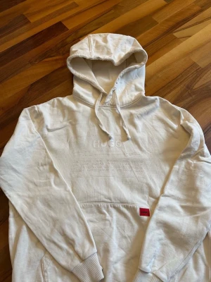 Vit hoodie från Hugo Boss med broderi - Säljer en stilren vit hoodie från Hugo Boss med broderad text på bröstet och en röd label vid fickan. Hoodien har klassisk huva med dragsko, magficka och ribbade muddar. Tillverkad i mjuk bomull för skön känsla.