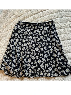 Svart blommig kjol från H&M Divided - Säljer en svart kortkjol från H&M Divided med ett härligt blommigt mönster av vita och gula prästkragar. Kjolen har en lätt utsvängd form och är gjord i ett mjukt syntetmaterial som känns bekvämt och luftigt. Perfekt för dig som gillar en lekfull och trendig stil.