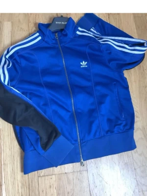 Blå Adidas ziptröja med vita ränder - Snygg blå ziptröja från Adidas med klassiska vita ränder längs ärmarna och Adidas-logga på bröstet. Tröjan har hög krage, dragkedja framtill och muddar vid ärmslut och nederkant. Materialet är glansigt och sportigt, perfekt för en streetwear-look.