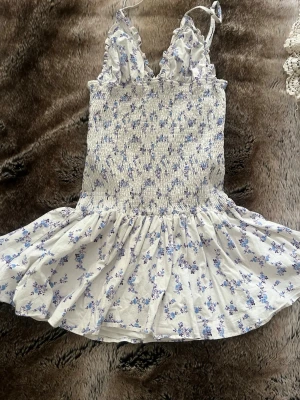 Blommig kortklänning med volang - Söt kortklänning med smala axelband och volangdetaljer vid bysten. Klänningen är vit med blått blommönster och har en smockad överdel som ger en bekväm passform. Kjoldelen är lätt utställd. perfekt för en sommarkväll