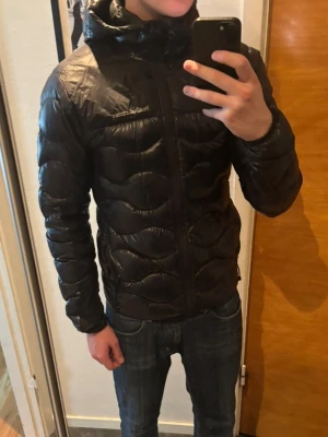 Svart dunjacka från Peak Performance - Snygg svart dunjacka från Peak Performance med huva och dragkedja framtill. Jackan har ett vågigt quiltat mönster och diskret logga på bröstet. Perfekt för kalla vinterdagar och riktigt lätt att bära. Klassisk passform och sportig look.