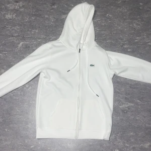 Vit hoodie från Lacoste - Säljer en stilren vit hoodie från Lacoste med dragkedja och klassisk krokodil-logga på bröstet. Den har en justerbar huva med snören och praktiska fickor framtill. Perfekt för en avslappnad look.