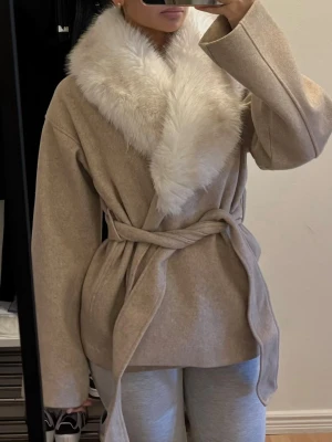 Beige kappa med fuskpäls - Stilren beige kappa med bred krage i vit fuskpäls och knytskärp i midjan. Kappan har en rak passform och vida ärmar, vilket ger en trendig och avslappnad look. Perfekt för dig som vill ha en elegant och mysig jacka till hösten eller vintern.