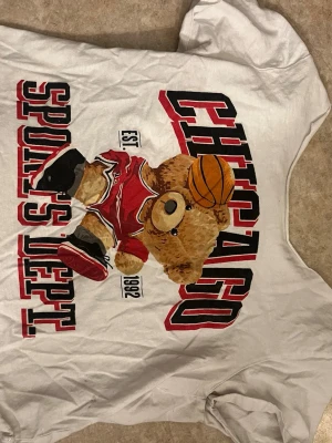 Vit Chicago t-shirt med björntryck - Säljer en vit t-shirt med ett stort tryck av en basketspelande björn i röd Chicago-uniform och texten 'Chicago Sports Dept'. T-shirten har korta ärmar och en avslappnad passform. Perfekt för dig som gillar sportiga och lekfulla motiv.