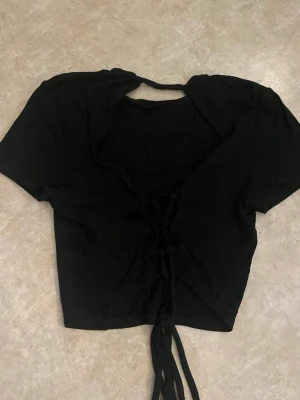 Svart croppad topp med snörning - Snygg svart croppad topp med korta ärmar och snörning framtill. Toppen har en öppen detalj i ryggen och är tillverkad i ett mjukt, stretchigt material. Perfekt för dig som gillar trendiga och edgy plagg.