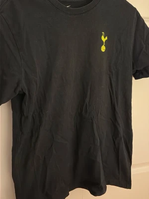 Svart Tottenham Hotspur t-shirt - Svart t-shirt från Tottenham Hotspur med klubbens gula logga tryckt på bröstet. Klassisk rund hals och korta ärmar. Tillverkad i mjuk bomull för en skön och avslappnad stil.