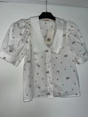 Vit blus med puffärm och spetskrage - Söt vit blus med små blommiga detaljer och stor spetskrage med broderi. Puffiga trekvartsärmar ger en trendig siluett. Knäpps framtill med knappar. Perfekt för dig som gillar romantisk och vintageinspirerad stil.