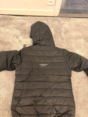 Svart dunjacka med huva Swedmount - Svart dunjacka från Swedmount med huva och dragkedja framtill. Jackan har två sidofickor och quiltad design med horisontella sömmar. Perfekt för kalla vinterdagar och har en stilren look som passar till det mesta.