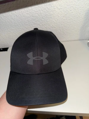 Svart keps från Under Armour - Snygg svart keps från Under Armour med det klassiska logotrycket framtill. Kepsen har böjd skärm och är tillverkad i ett lätt och slitstarkt material. Insidan har svettband med texten 'Blitzing'. Perfekt för en sportig och avslappnad stil.