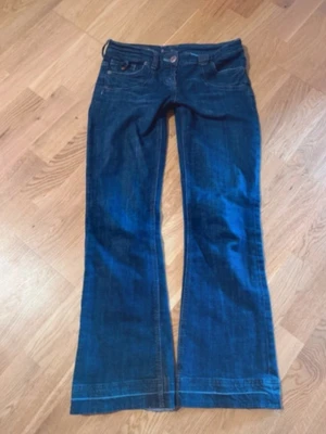 Blå lågmidjade bootcut jeans från Replay - Säljer ett par mörkblå bootcut jeans från Replay med snygga kontrastsömmar och detaljerade bakfickor. Jeansen har låg midja och ett coolt patch på ena bakfickan. Perfekta för dig som gillar retrovibbar och unik design.