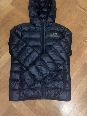 EA7 jacka  - Snygg svart dunjacka från EA7 Emporio Armani med huva och dragkedja framtill. Jackan har ett diskret EA7-logotyptryck på bröstet och är quiltad för extra värme. Perfekt till våren och har en modern, sportig look. Kom gärna med frågor. Äkta EA7 jacka. Passar 160-172. Bra skick, kommer med en liten påse man kan packa jackan vilket gör det väldigt enkelt att ta med den utan att behöva ha på sig den. Finns en gucciväska också men kan inte lägga upp den, kom pv för frågor och intresse om den. 