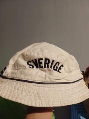 Vit bucket hat Sverige Retro - Vit bucket hat från Retro med svart broderad text 'Sverige' framtill och en liten svensk flagga på sidan. Fiskehatt modell. Obs!! Gosedjuren, dinosaurien på sista bilden medföljer EJ, dino är enbart en modell för att enkelt visa upp hatten!!