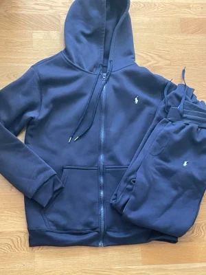 Mörkblå loungewear-set från Polo Ralph Lauren - Mysigt mörkblått loungewear-set från Polo Ralph Lauren med hoodie och matchande byxor. Hoodien har dragkedja, fickor och dragsko i huvan. Både tröja och byxor har den klassiska Polo-loggan broderad i vitt. Perfekt för chill dagar hemma.