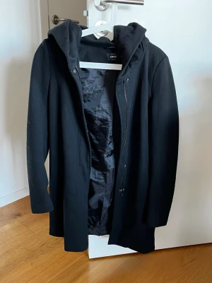 ONLY svart kappa med huva stl S - En tidlös svart kappa från märket ONLY. Kappan har en stor, mysig huva. Dragkedja framtill, tryckknappar och en rymlig huva. Fodrad på insidan.  Kappan är i fint använt skick, men det förekommer en del ytlig nopprighet/ludd på materialet (se närbild). Går säkert att fräscha upp med en noppmaskin