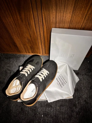 Svarta sneakers från Maison Margiela - Svarta sneakers från Maison Margiela med klassisk låg siluett, vita snören och gummisula i brunt. Ovandelen är i läder och mocka, och insidan är fodrad i ljust material. Snygg minimalistisk design med diskreta detaljer.