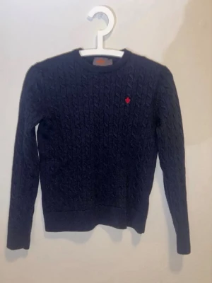 Mörkblå kabelstickad tröja - Mörkblå kabelstickad tröja med rund halsringning och ribbade muddar. Liten röd broderad detalj på bröstet. Perfekt för lager på lager och en klassisk look. Tröjan har lång ärm och är gjord i mjukt material.