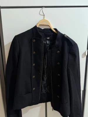 Svart kavajjacka från H&M med knappar - Stilren svart kavajjacka från H&M med dekorativa guldknappar framtill och axelklaffar. Jackan har rak passform, lång ärm och dragkedja. Materialet är en blandning av polyester och ull som ger en klassisk känsla.