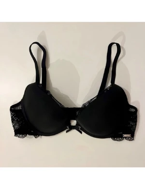 Svart ella m push-up bh med spetsdetaljer - Snygg svart push-up bh med vadderade kupor och fina spetsdetaljer längs kanterna. Justerbara axelband och liten rosett framtill. 