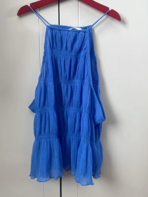 Blå volangig linnetopp - Säljer en blå linnetopp med tunna axelband och volangig, luftig design. Toppen har en rynkad struktur och är perfekt för varma dagar. Passar dig som gillar en avslappnad och trendig stil.