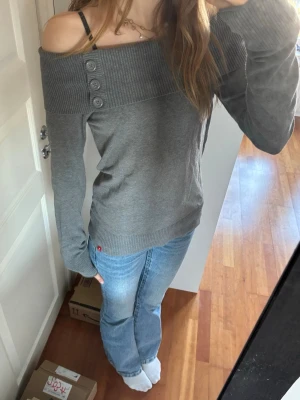Grå off shoulder stickad tröja - Trendig grå stickad tröja med bred ribbad off shoulder-krage och tre dekorativa knappar på axeln. Långärmad modell med ribbade muddar vid ärmslut och nederkant.🤍