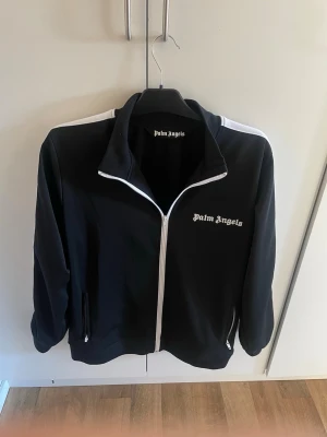 Svart zip-hoodie från Palm Angels - Svart hoodie från Palm Angels med vit dragkedja och vita detaljer längs axlarna. Klassisk logga på bröstet och två fickor med dragkedja. Snygg och sportig design som passar perfekt till streetwear-stilen.