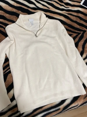 Vit ribbstickad tröja med halv dragkedja - Snygg vit ribbstickad tröja med halv dragkedja och hög krage. Tröjan har långa ärmar och är tillverkad i ett mjukt, stretchigt material som känns skönt mot huden. Perfekt för lager på lager och enkel att matcha med olika outfits.