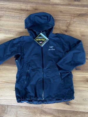 Svart vindjacka från Arc'teryx - Snygg svart vindjacka från Arc'teryx med huva och dragkedja framtill. Tillverkad i slitstarkt GORE-TEX-material som står emot både vind och regn. Två praktiska fickor med dragkedja och justerbara muddar. Perfekt för dig som vill ha en lätt och funktionell jacka.