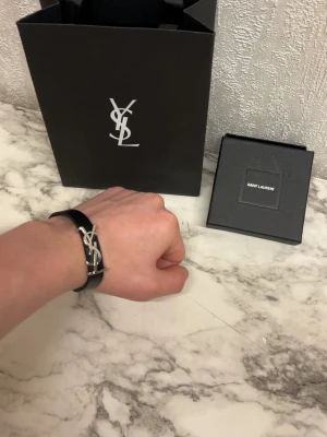 Saint Laurent svart läderarmband - Snyggt svart läderarmband från Saint Laurent med ikoniskt YSL-logo i silverfärgad metall. Armbandet har ett elegant spänne där det står 'Saint Laurent Paris'. Perfekt accessoar för att lyfta din stil.