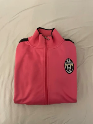 Juventus rosa träningsjacka - Snygg rosa träningsjacka från Juventus med svart logga på bröstet och svarta detaljer längs axlar och ärmslut. Jackan har hel dragkedja, ståkrage och två fickor med dragkedja. Perfekt för dig som vill sticka ut på träningen eller på stan.