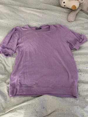 Ljuslila topp med puffärm - Säljer en ljuslila topp med puffärmar och rund halsringning. Toppen har en avslappnad passform och är gjord i mjukt bomullsliknande material. Perfekt för dig som gillar en enkel men trendig stil.